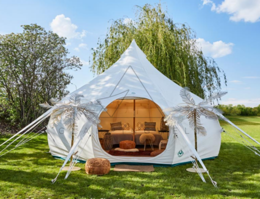 A white, spacious glamping tent with decorative elements is set up in a grassy area with trees and clear blue sky in the background. The interior of the tent is visible.